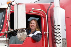 photo of a caucasian female truck driver sitting in the driver seat of her red semi truck, smiling towards the camera.