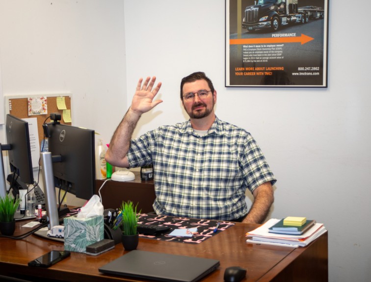 A smiling staff member in glasses and a plaid shirt waves from behind his office desk, with dual monitors beside him