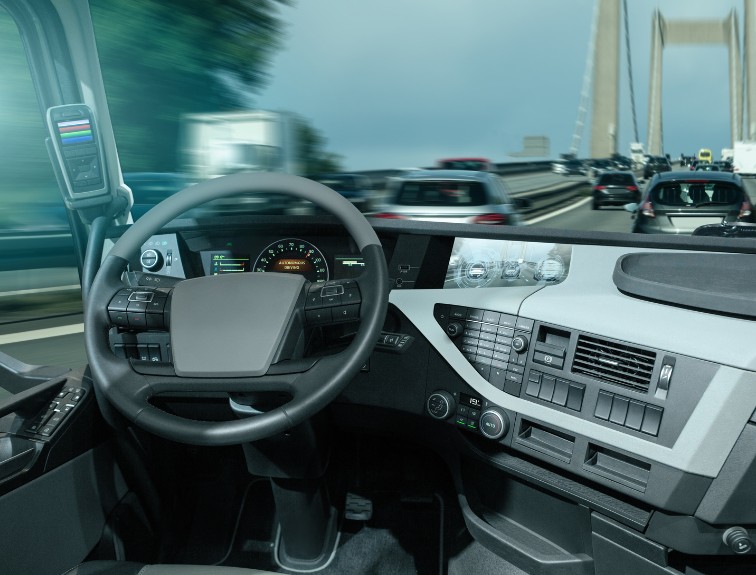 View from inside the cab of a self-driving semi-truck showing the steering wheel, digital dashboard displays, and windshield looking out at highway traffic