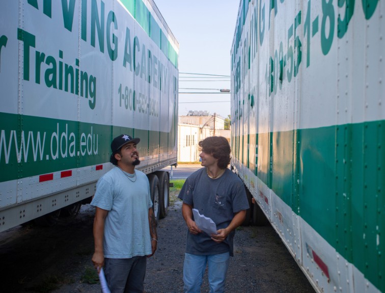Two DDA students holding papers talk between two parked trailers on a sunny day.
