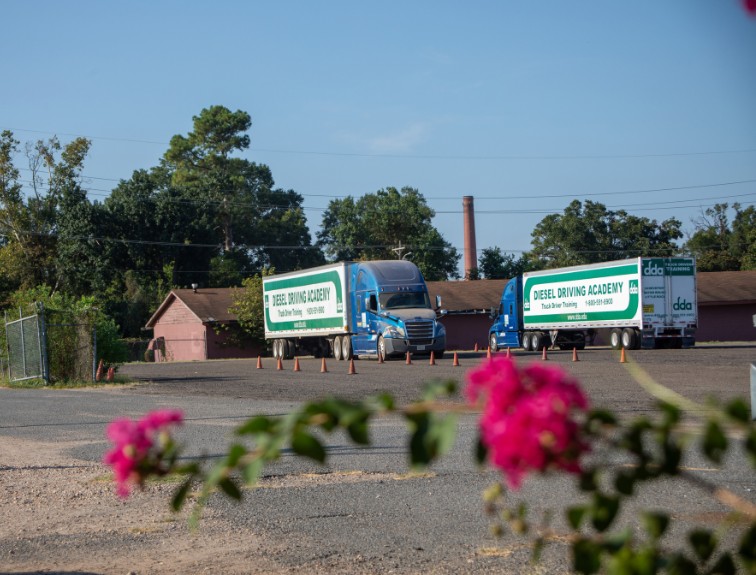 Two blue DDA semi trucks with green and white trailers sit on a training yard lined with orange cones, framed by pink flowers in the foreground.