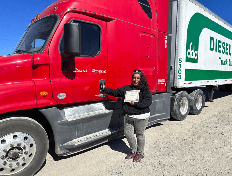 Image of female truck driver who is holding her CDL training certificate and standing with one hand on the red school trailer