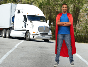 Image of male truck driver with blue shirt, blue pants, and gray sneakers, with a red cape around his neck, standing in front of a white American semi truck.